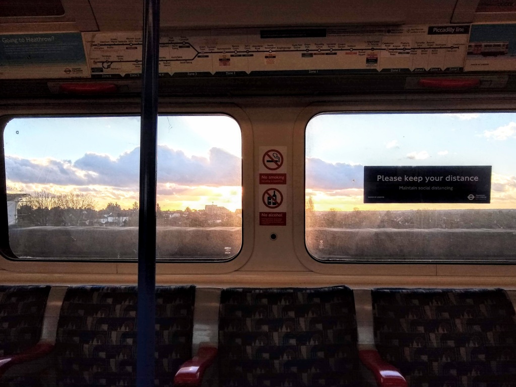 In a Piccadilly Line train on the raised line west from Hammersmith, looking across at the windows opposite, no one seated there. Outside there is a low glaring sun flare in blue sky behind the roofs of buildings in the distance. Among the official signs and adverts, there is an official London Underground sign on a window that says “Keep your distance. Maintain social distancing.” (February, 2022)